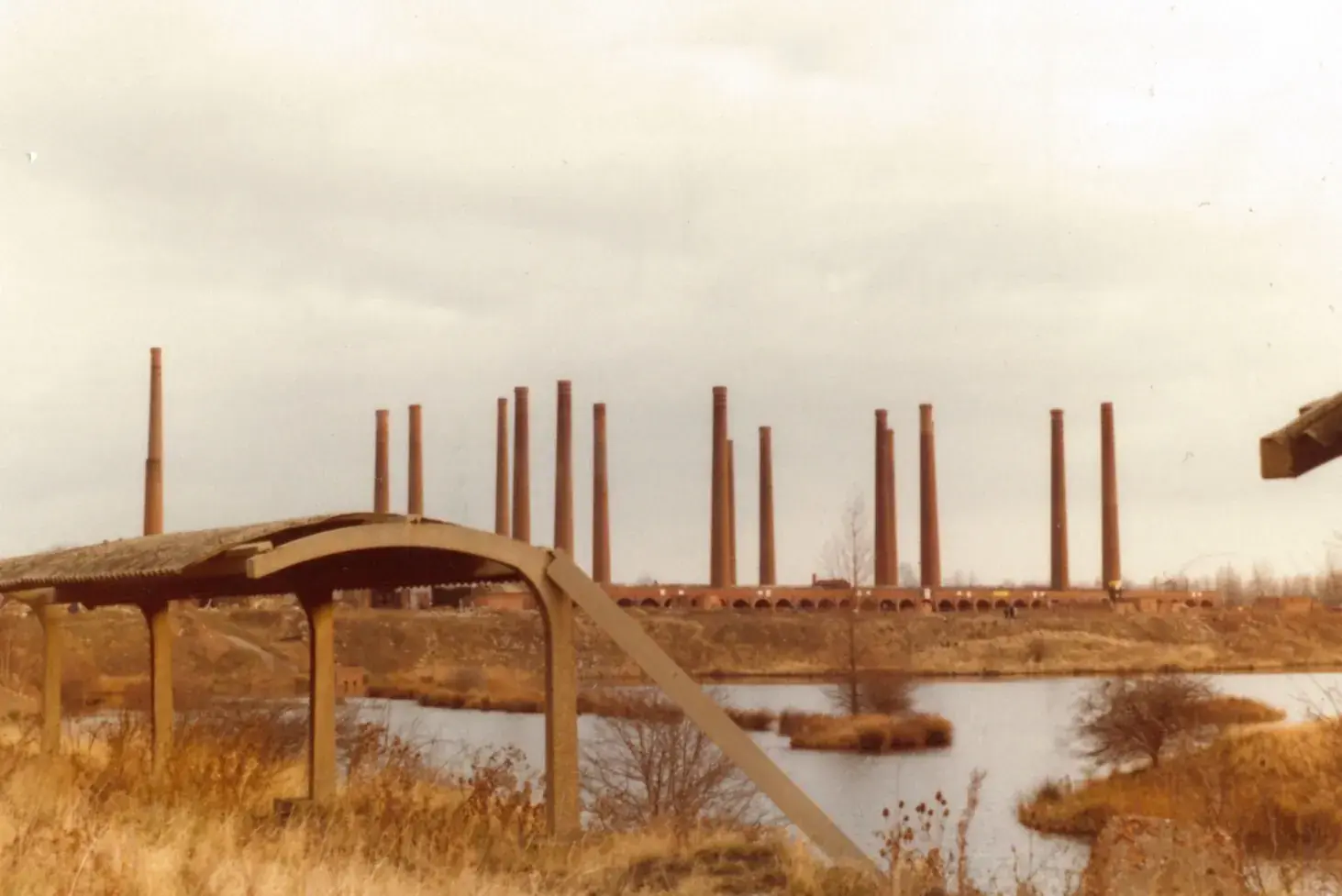 A row of tall brick chimneys on the skyline beyond flooded clay pits at the old brickworks