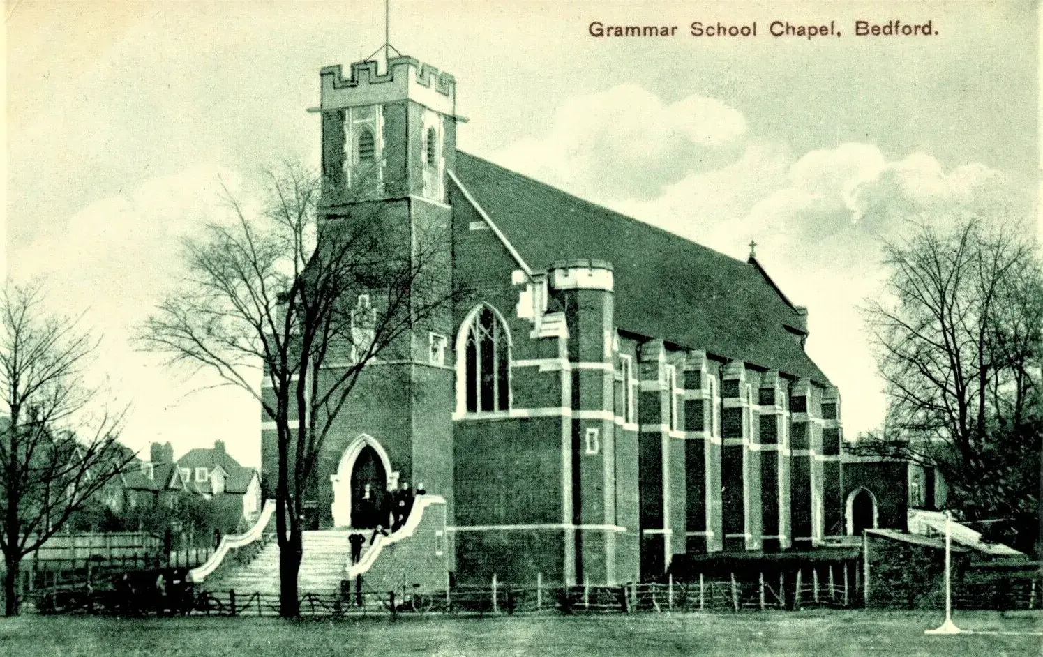 A brick chapel with a tower, arched windows, and steps leading to the entrance, surrounded by trees and a grassy area